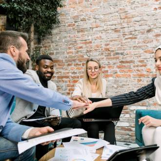 Successful multiethnic team of smart business people celebrating success of a new project and putting hands palms together in the center above the table at modern loft office.